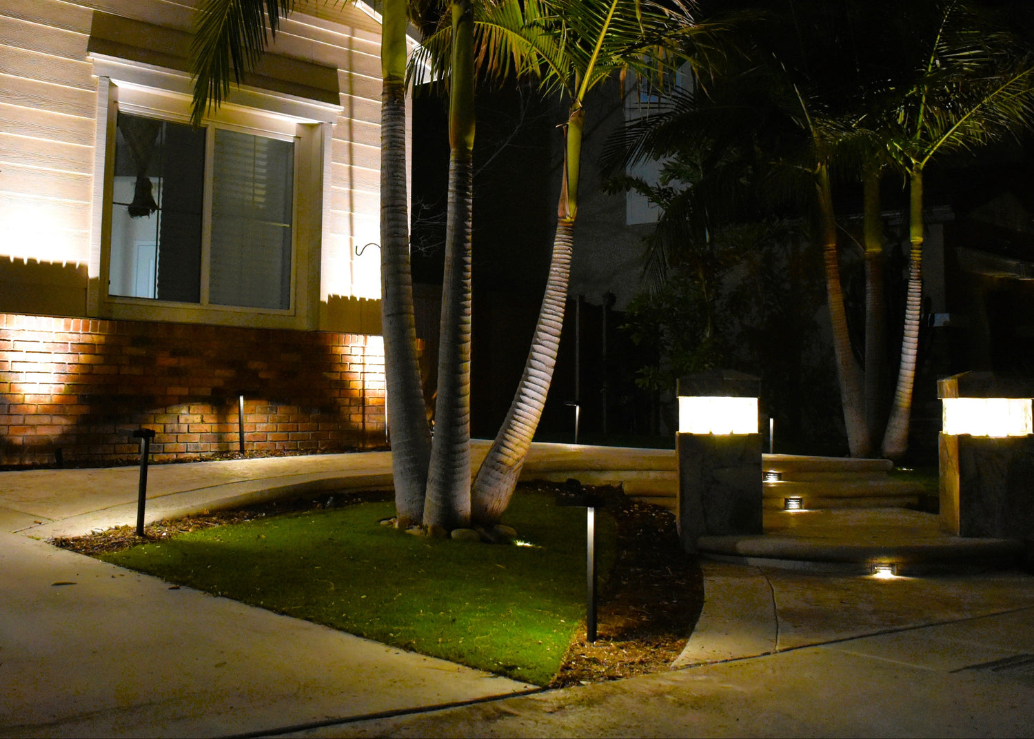Outdoor landscape lighting at night showing a house exterior, palm trees, walkways, grass, and modern rectangular bollard and ground-mounted lights.