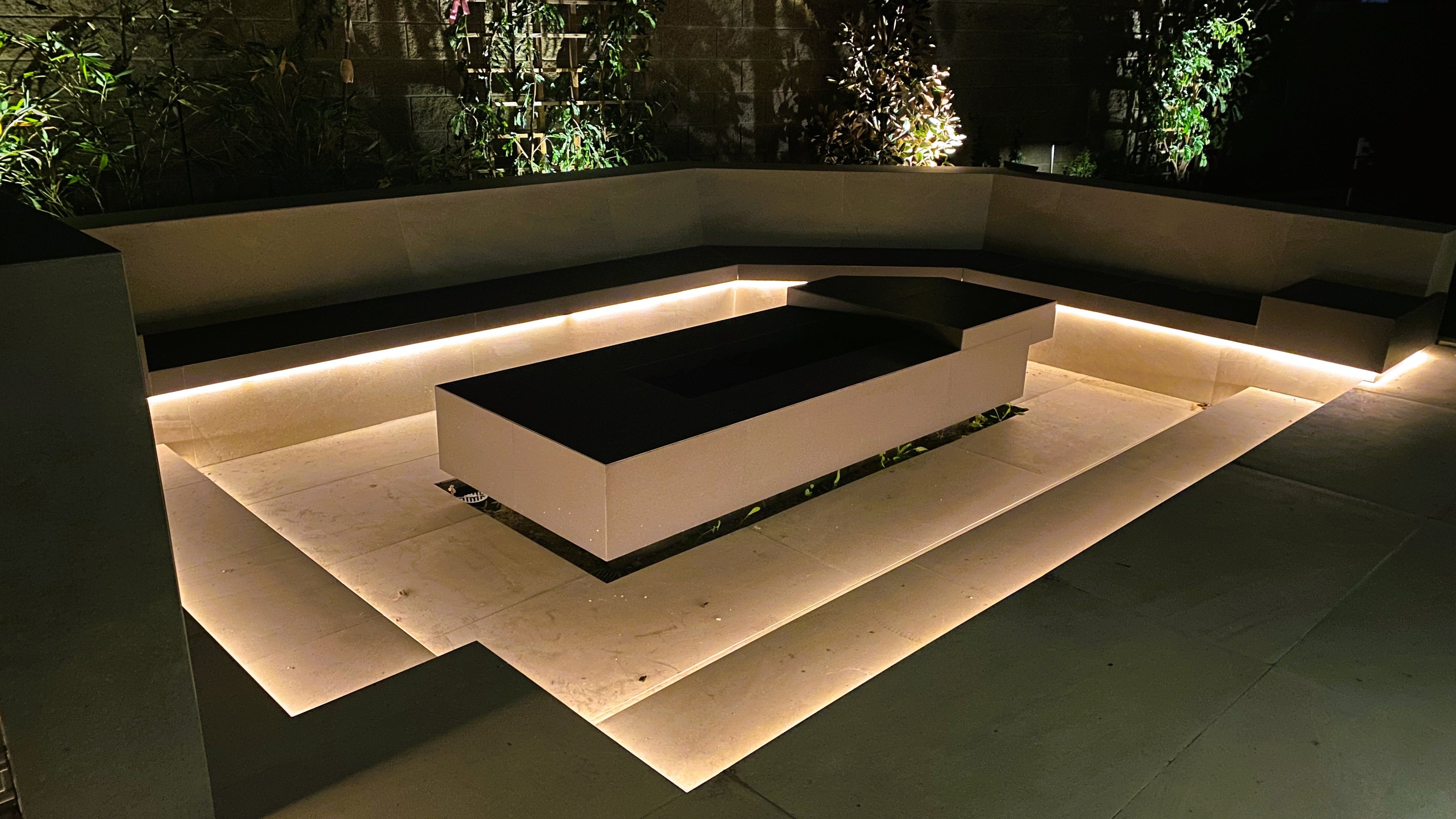 Modern outdoor patio seating area at night with a rectangular black bench or fire pit centered on a raised light stone platform, surrounded by warm under-step lighting and dark greenery in the background.