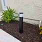 A black cylindrical outdoor bollard light with a frosted white top, installed in a mulched garden bed next to a stucco house wall.
