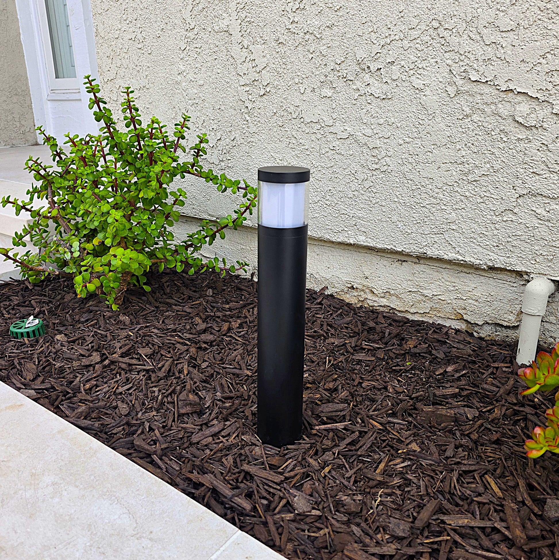 A black cylindrical outdoor bollard light with a frosted white top, installed in a mulched garden bed next to a stucco house wall.