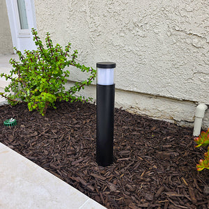 A black cylindrical outdoor bollard light with a frosted white top, installed in a mulched garden bed next to a stucco house wall.