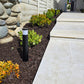 A black outdoor bollard light with a white cylindrical sensor band, installed in a landscaped garden bed next to a light stone patio walkway and steps.