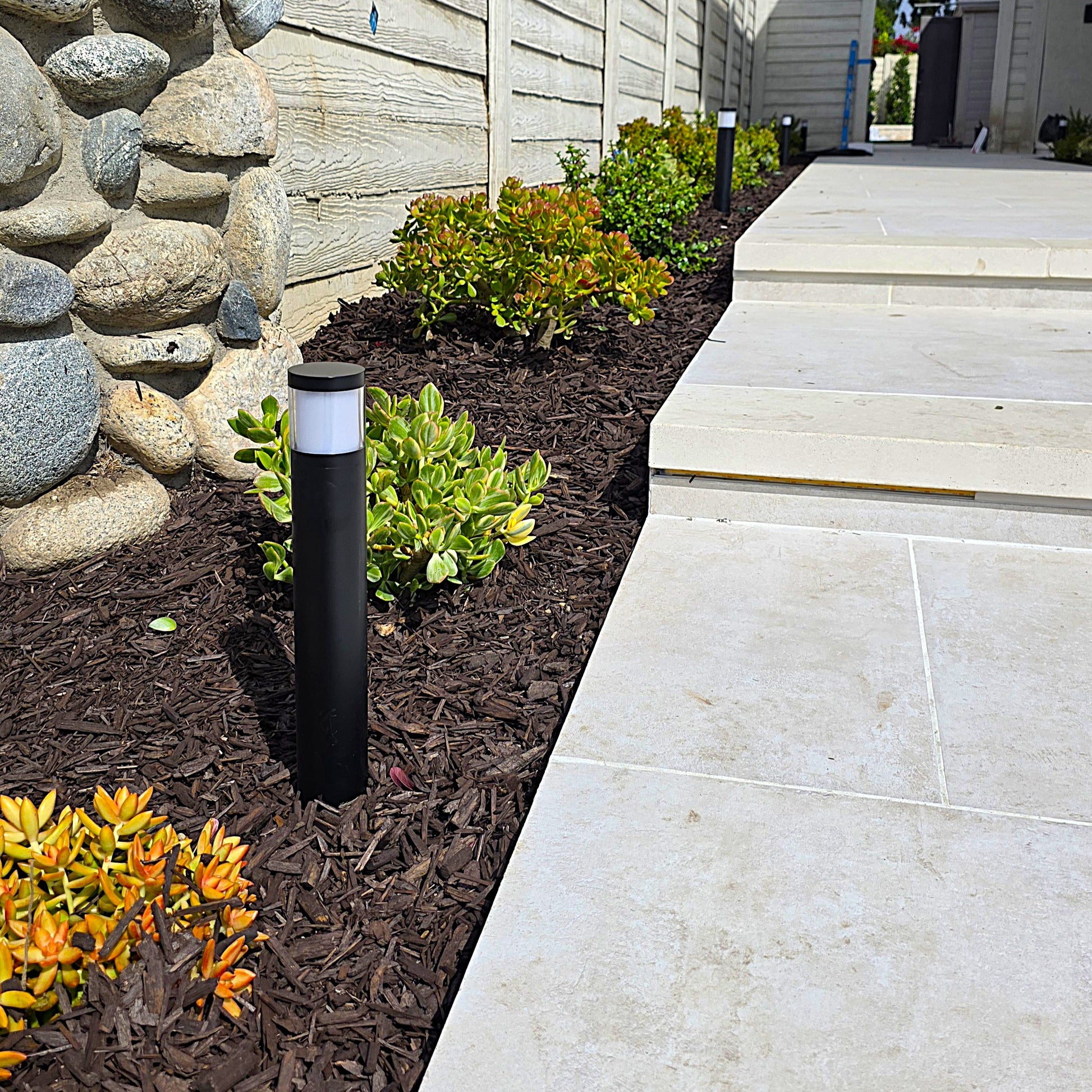 A black outdoor bollard light with a white cylindrical sensor band, installed in a landscaped garden bed next to a light stone patio walkway and steps.