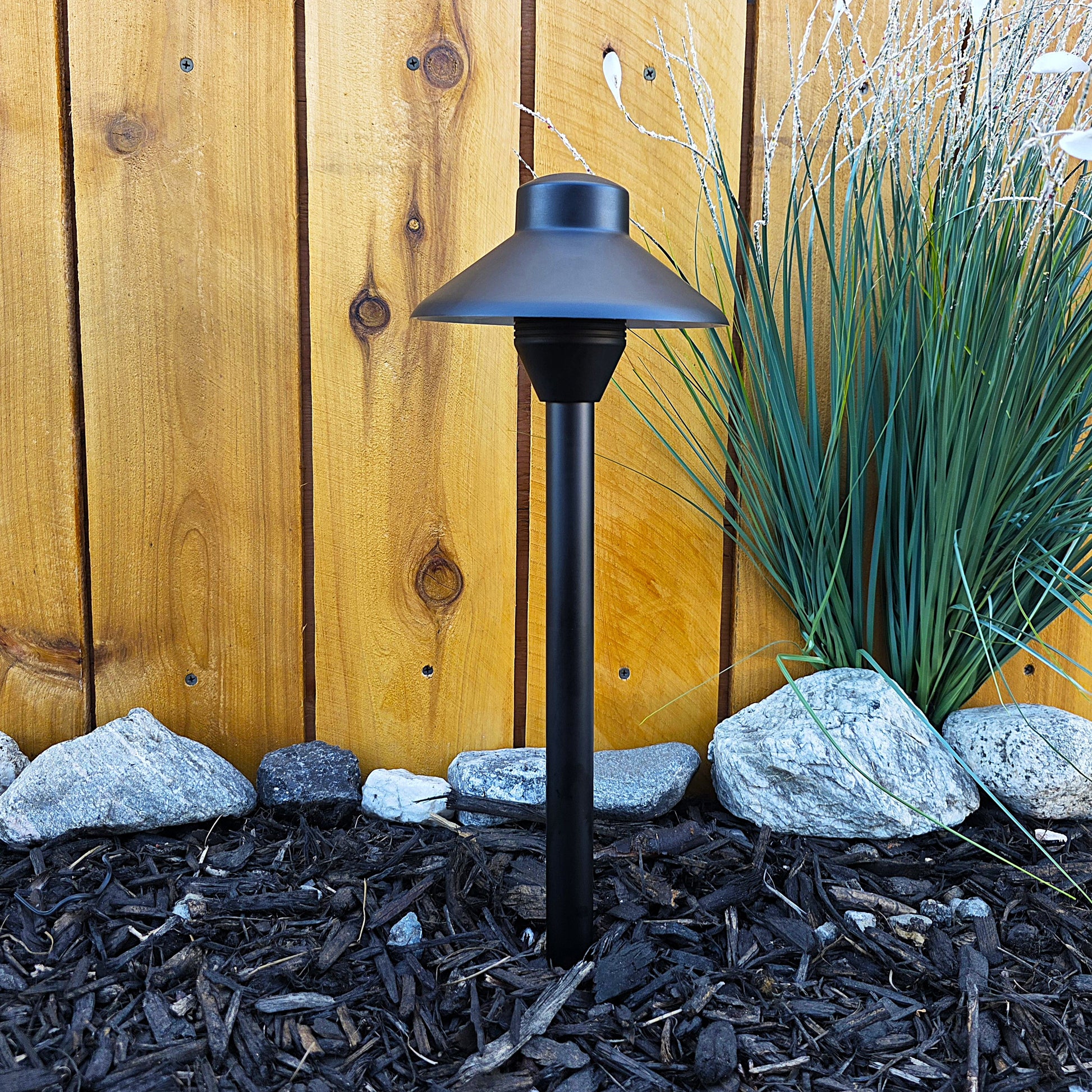 Cascade Cast Brass Path Light outdoor landscape lighting on a post beside a wooden fence and rocks
