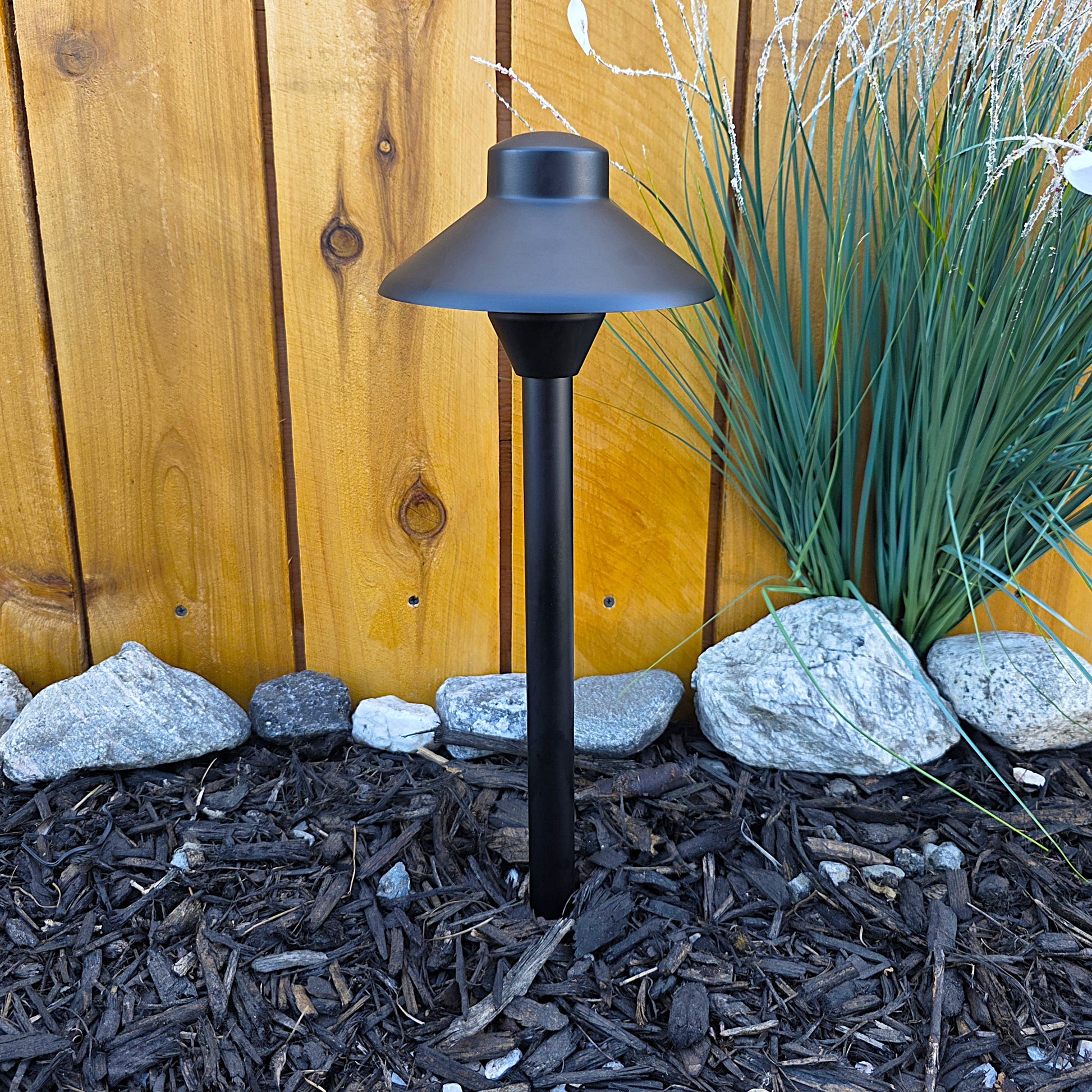 Cascade Cast Brass Path Light outdoor landscape fixture on a garden path beside mulch and rocks near a wooden fence.