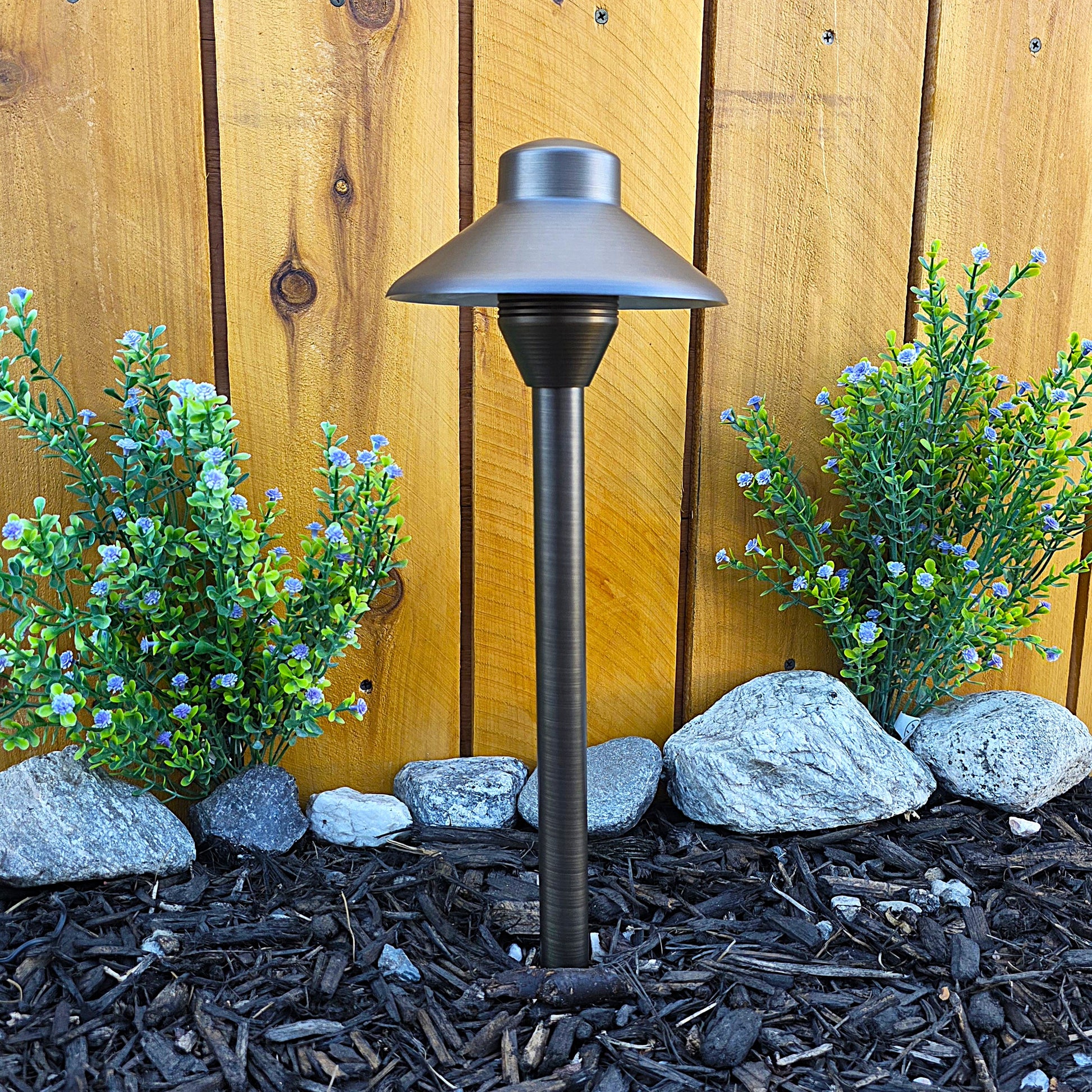 Cascade Cast Brass Path Light, solid brass outdoor landscape lighting for garden paths beside shrubs and wood fence.