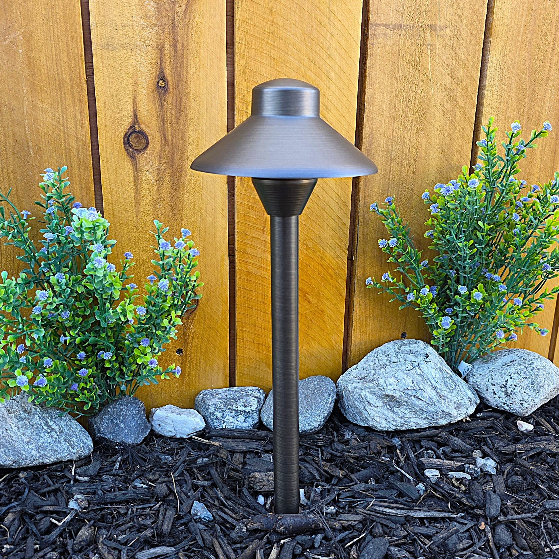 Cascade Cast Brass Path Light outdoor landscape lighting along walkway beside a wooden fence with blue flowers.