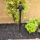 Black outdoor lamp post in a garden setting with plants and a brick wall.
