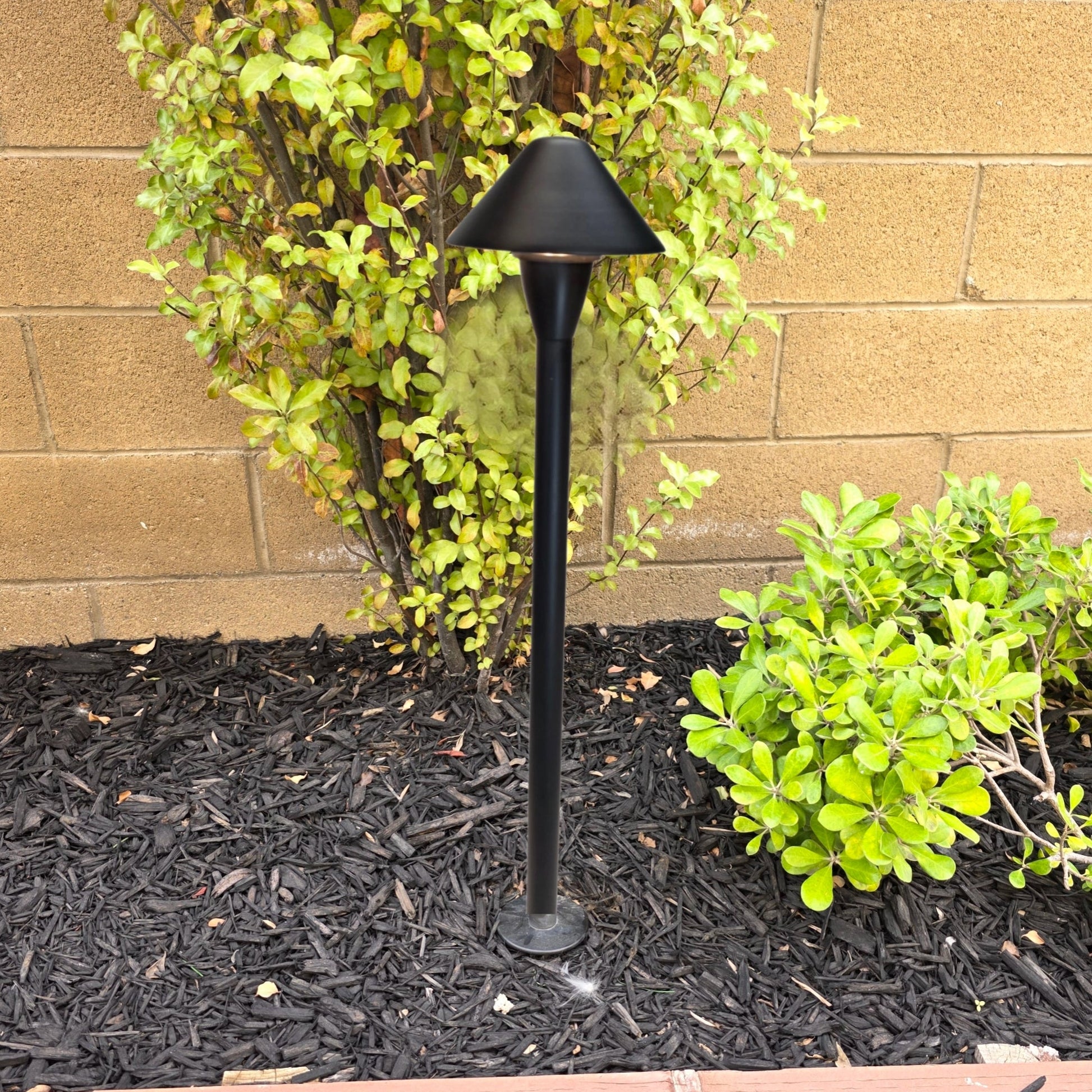 Black outdoor lamp post in a garden setting with plants and a brick wall.