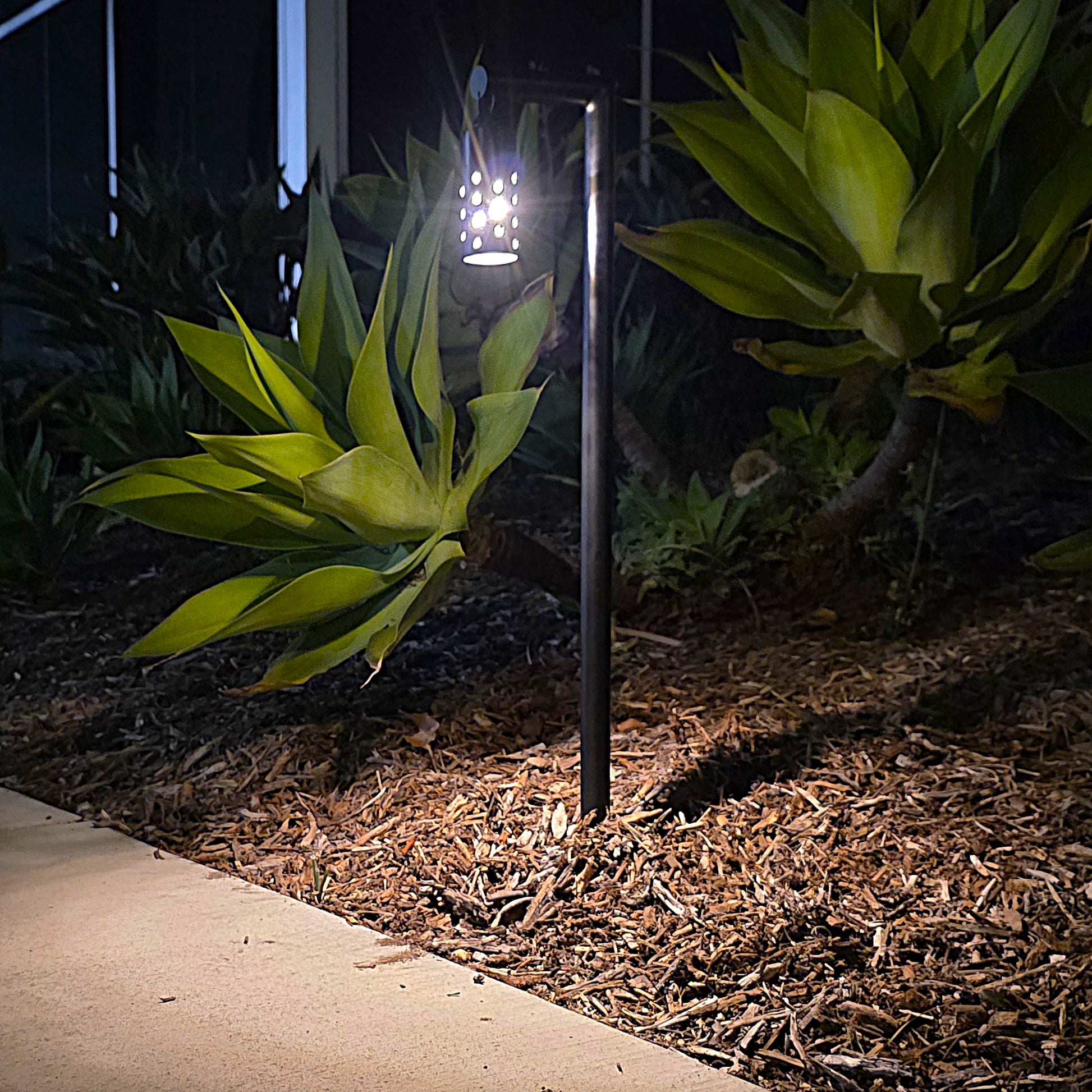 Pendule solid cast brass black lantern path light in outdoor landscape, casting warm LED glow on mulch and plants.